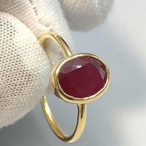May include: A gold ring featuring a large, oval-cut ruby gemstone. The ruby is a deep red color and is set in a gold bezel. The ring's band is a simple, polished gold.