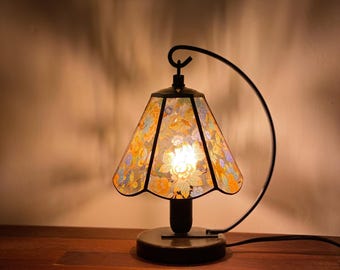 Può includere: Lampada da tavolo decorativa con paralume a motivi floreali nei toni del giallo, arancione e blu. La lampada ha una montatura e una base in metallo nero e un braccio curvo. La lampada è accesa, illuminando il motivo floreale.