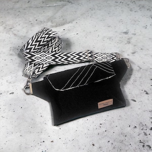 May include: A black crossbody bag with a geometric patterned strap in black and white. The bag has a flap closure with white stitching and a small rectangular tag. The strap is adjustable and attached with silver-tone hardware.