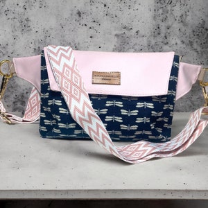 May include: A pink and navy blue fanny pack with a patterned strap. The bag features a pink flap and a navy blue body with a dragonfly pattern. The strap is white with a pink geometric design. Gold-colored hardware.