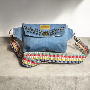 May include: Light blue denim fanny pack with a studded flap and a colorful patterned strap. The bag features a gold-tone clasp and a small rectangular label. The strap is a woven design with red, yellow, blue, and green accents.