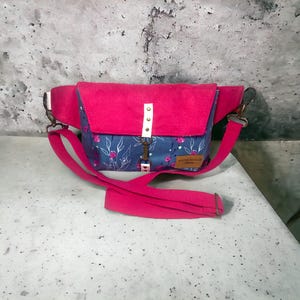 May include: A pink and blue floral fanny pack with a white strap and a metal clasp. The bag has a pink flap with white dots and a small brown leather tag with the text "Something Remarkable" on it.