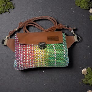 May include: A colorful woven fanny pack with a brown leather flap and strap. The pack features a rainbow pattern of red, yellow, green, and blue. It has a silver clasp and a small brown label.