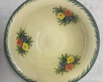 May include: A round, cream-coloured ceramic serving bowl with a green, textured rim. The bowl features three clusters of painted flowers in red, yellow, and blue. The flowers are surrounded by green leaves.
