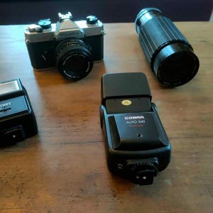 May include: A selection of vintage photographic equipment, including a black and silver Fujica camera with a lens, a black telephoto lens, and two black flash units. The flash units are labelled Unomat 200 and Cobra Auto 260.