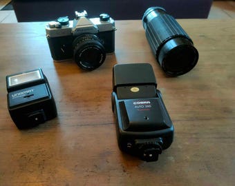 Könnte beinhalten: Eine Sammlung von Vintage-Fotoausrüstung, darunter eine schwarz-silberne Fujica-Kamera mit Objektiv, ein schwarzes Teleobjektiv und zwei schwarze Blitzgeräte. Die Blitzgeräte sind mit Unomat 200 und Cobra Auto 260 beschriftet.