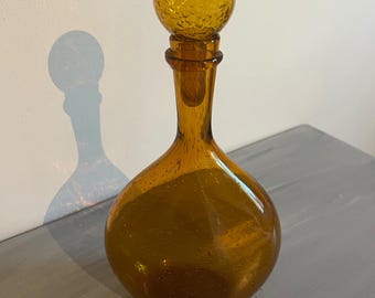 May include: Amber glass decanter with a spherical body and a slender neck. The stopper is a textured sphere. The decanter is on a grey surface, with a shadow on the white wall. The glass has small bubbles.