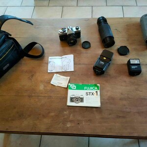May include: A selection of vintage camera equipment, including a camera body, lenses, flashes, lens caps, and a camera bag. The camera is silver and black, and the bag is black with yellow accents. The equipment is arranged on a wooden surface.