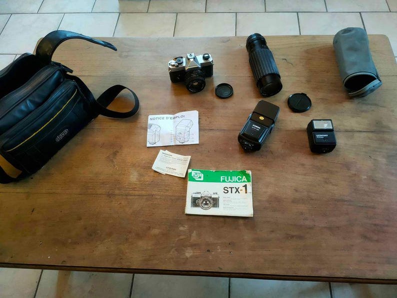 May include: A selection of vintage camera equipment, including a camera body, lenses, flashes, lens caps, and a camera bag. The camera is silver and black, and the bag is black with yellow accents. The equipment is arranged on a wooden surface.