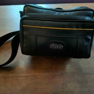 May include: Black rectangular camera bag with a shoulder strap. The bag has a gold stripe and the brand name "daxa" in a white oval. The bag is on a wooden surface.