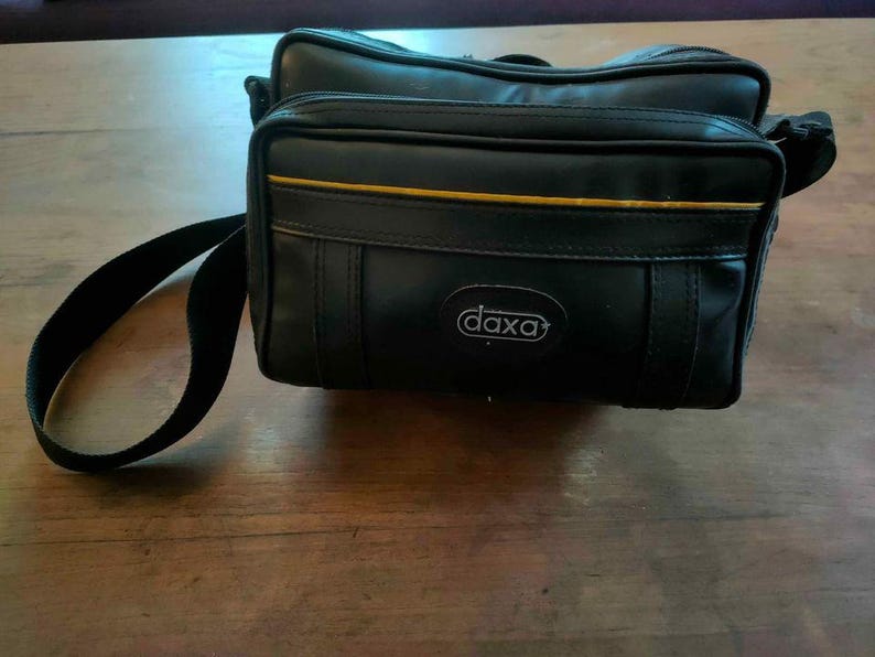May include: Black rectangular camera bag with a shoulder strap. The bag has a gold stripe and the brand name "daxa" in a white oval. The bag is on a wooden surface.