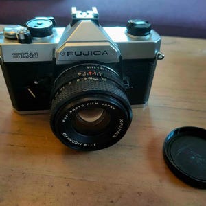 May include: A Fujica STX-1 35mm film camera with a black body and silver top. The lens is a Fujinon 50mm f/1.9. A black lens cap sits to the right. The camera is on a wooden surface.