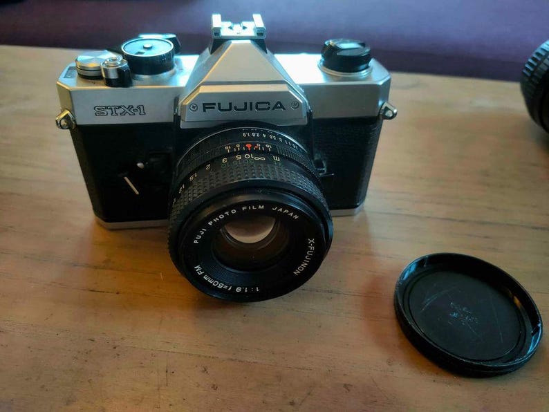May include: A Fujica STX-1 35mm film camera with a black body and silver top. The lens is a Fujinon 50mm f/1.9. A black lens cap sits to the right. The camera is on a wooden surface.