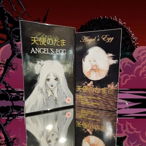 May include: Two VHS tapes with the title "Angel's Egg" in English and Japanese. The cover art features a young woman with long white hair and a white dress. The background is a dark, abstract design.