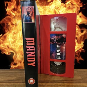 Könnte beinhalten: Ein rotes VHS-Kassettengehäuse mit dem Titel "MANDY" in weiß auf der Vorderseite. Das Gehäuse ist geöffnet und zeigt die VHS-Kassette im Inneren. Die Kassette ist schwarz mit einem roten Etikett, auf dem "MANDY" in weiß steht. Das Gehäuse steht auf einer Holzoberfläche.