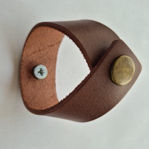 May include: A brown leather loop handle featuring a smooth outer surface and a textured inner side. The handle is secured with a circular brass-toned metal fastener on the front and a silver-toned Phillips head screw visible on the inner side.
