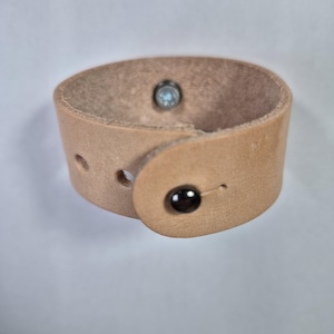 May include: A tan leather cuff bracelet with a wrap-around design. The bracelet features a black button closure and a silver-colored rivet. The leather has a natural, textured appearance, and the bracelet is approximately 1.5 inches wide.