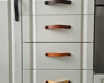 Può includere: Un set di cinque maniglie per cassetti in pelle fissate su ante e cassetti di armadi bianchi. Le maniglie sono disposte in una sfumatura di colore verticale, che va dal marrone scuro in alto al beige chiaro in basso, fissate con viti nere.