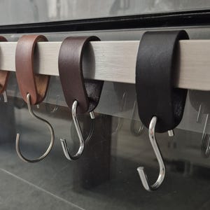May include: Close-up of a row of leather S-hooks in varying shades of brown and black, hanging from a silver bar. The hooks are made of leather and have a polished metal finish.
