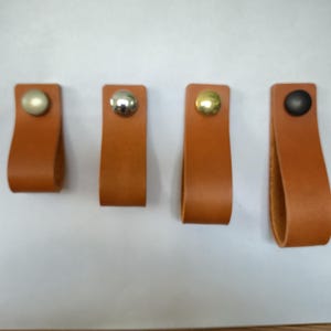 May include: Four brown leather cabinet pulls with metal rivets. The rivets are silver, gold, and black. The pulls are rectangular with a loop at the bottom, designed for easy gripping. The leather has a smooth texture.