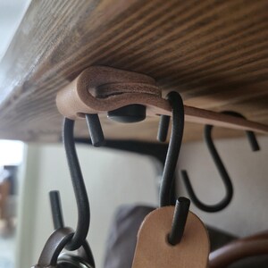 Peut inclure: Une sangle en cuir beige clair est pliée et fixée avec une attache noire sous une étagère en bois. Plusieurs crochets en S en métal noir mat sont suspendus à la sangle en cuir, offrant une solution de rangement suspendue pour divers objets.