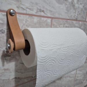 May include: A wall-mounted paper towel holder. The holder features a light brown leather strap and a wooden dowel to hold the paper towels. The paper towel roll is white with a repeating circular pattern. The holder is attached to a tiled wall.