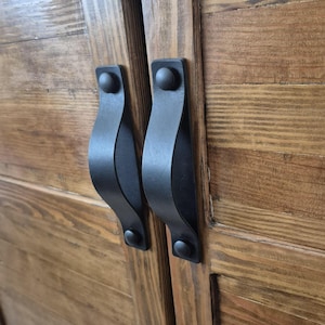Pode incluir: Dois puxadores de gaveta em metal preto mate com um design curvo e arqueado, cada um fixado por duas cabeças de parafuso circulares. Os puxadores estão montados verticalmente num par de portas de armário de madeira com grão castanho.