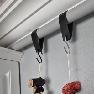 Peut inclure: Deux lanières en cuir noir pliées sur une tige horizontale, chacune fixée par un crochet en S en métal. Un crochet est noir et l'autre est argenté. Des aliments séchés sont suspendus aux crochets par de fines ficelles blanches.