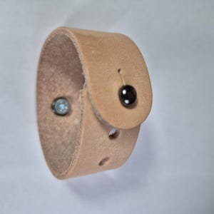 Peut inclure: Un bracelet manchette en cuir marron clair avec une fermeture à bouton. Le bracelet est orné d'un bouton marron foncé et d'un clou de couleur argentée. Le bracelet est conçu pour s'enrouler autour du poignet.