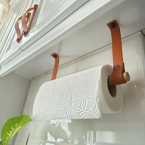 May include: A paper towel holder with a roll of white paper towels. The holder is made of brown leather straps and a wooden dowel. The holder is mounted on a white cabinet.