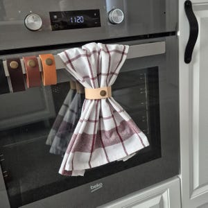 Può includere: Un canovaccio da cucina bianco con motivo a quadri rosso scuro appeso alla maniglia di un forno, fissato da un cinturino in pelle color cuoio chiaro con un bottone a pressione in metallo color ottone. Sono visibili tre cinturini in pelle aggiuntivi.