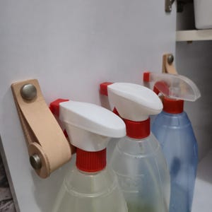 Peut inclure: Trois flacons pulvérisateurs avec des buses blanches et des colliers rouges. Les flacons transparents contiennent du liquide. Une sangle en cuir marron clair avec des rivets métalliques maintient les flacons en place.