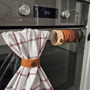 Può includere: Un canovaccio bianco con motivo a griglia rosso scuro tenuto fermo da una cinghia in pelle color cuoio con un bottone a pressione in ottone. Una fila di cinque cinghie in pelle aggiuntive in varie tonalità di marrone e nero pende dal forno.