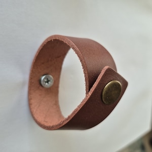 May include: A brown leather cuff bracelet featuring a smooth exterior and a raw, textured interior edge. The bracelet is secured with a metallic, circular brass-toned snap fastener on the front and a silver-toned screw head on the side.