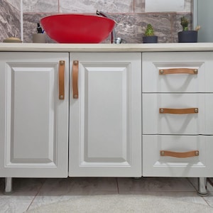 May include: A white bathroom vanity cabinet featuring two doors and three drawers, all equipped with tan leather strap handles. The vanity has a light-colored countertop with a large, round, bright red vessel sink and a chrome faucet fixture.