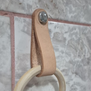 May include: A light brown leather towel holder with a natural wood ring. The leather strap is attached to the wall with a silver screw. The towel ring is made of light-colored wood. The holder is mounted on a marble tile wall.