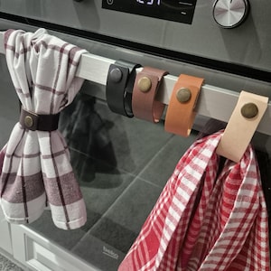 May include: A close-up of a kitchen appliance with a metal bar holding several leather towel holders. The holders are in black, brown, and tan, each with a metal snap. Two checkered kitchen towels are attached.