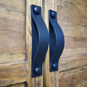 May include: Close-up of two black leather cabinet pulls attached to a wooden cabinet door. The pulls are curved and secured with black rivets. The wood has a natural grain and a warm, brown tone.