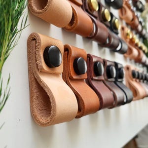 May include: A collection of leather loops arranged in rows, each featuring a folded design secured with a circular black or metallic snap button. The loops vary in shades of tan, brown, and black, creating a repetitive pattern of functional hardware.