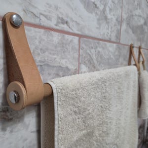 Könnte beinhalten: Ein Wandhandtuchhalter mit einem hellbraunen Lederriemen, der mit silberfarbenen Metallbeschlägen befestigt ist. Ein glatter Holzstab führt durch die Lederschlaufe und trägt ein strukturiertes, cremeweißes Handtuch. Ein zweiter, identischer Halter ist im Hintergrund zu sehen.