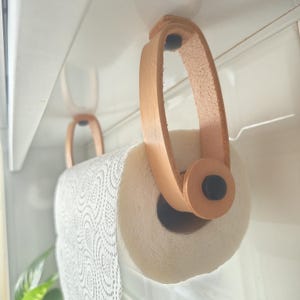Può includere: Un portarotolo da cucina con due cinghie in pelle color cuoio chiaro sospese a una superficie. Le cinghie sono fissate con chiusure circolari nere alle estremità e sostengono un rotolo di carta assorbente bianca con motivo a onde.