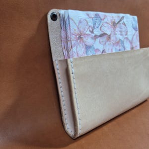 May include: A light tan leather napkin holder featuring white stitching along the edges and a single dark metal grommet at the top corner. The holder contains a stack of paper napkins decorated with a soft pink and white floral pattern.