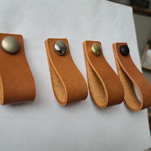 May include: Four leather drawer pulls in a warm tan color. Each pull features a loop design and is secured with a metal rivet. The rivets are silver, gold, and black. These pulls are suitable for cabinets and drawers.