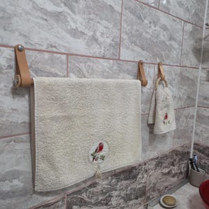 Peut inclure: Une serviette couleur crème avec un écusson brodé circulaire représentant un oiseau à poitrine rouge, suspendue à une barre porte-serviettes en cuir beige. Une serviette assortie plus petite pend à un anneau en bois fixé par une lanière en cuir.