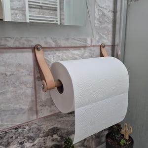 Peut inclure: Un porte-essuie-tout mural composé de deux lanières en cuir beige clair fixées par des éléments métalliques argentés. Une tige cylindrique en bois relie les lanières et soutient un rouleau d'essuie-tout blanc au motif en spirale texturé.