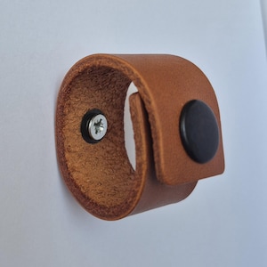 May include: A tan leather loop fastener featuring a smooth exterior and a textured interior surface. The loop is secured with a black circular snap button on the front and a silver-colored metal screw head visible on the inner side of the leather band.