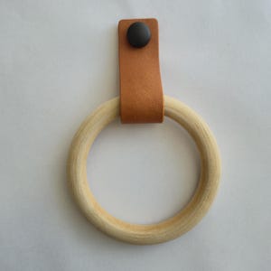 Peut inclure: Un anneau en bois de couleur claire fixé à une sangle en cuir beige. La sangle en cuir est repliée sur le dessus de l'anneau en bois et fixée par un seul bouton-pression en métal marron foncé, créant une boucle pour suspendre ou décorer.
