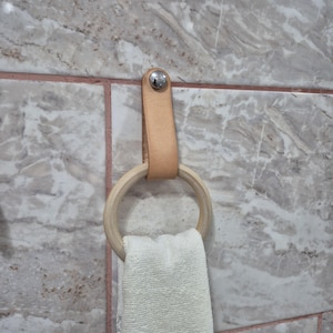 Può includere: Un portasciugamani in pelle colorata con un anello di legno e un piccolo asciugamano bianco. Il supporto è fissato al muro con un rivetto argentato. Lo sfondo è una parete di piastrelle di marmo.