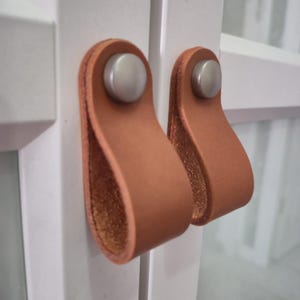 Op de afbeelding: Twee kastgrepen met een lichtbruine leren lus en een zilverkleurige metalen klinknagel. De grepen zijn gemonteerd op een witte kastdeur. De leren lussen zijn ongeveer 7.6 cm lang.
