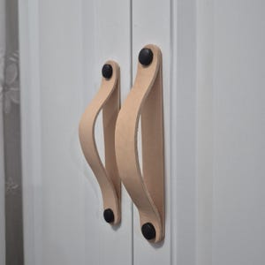 May include: Two light tan leather drawer pulls attached to a white cabinet door. Each handle is a curved strip of leather secured at both ends by small, circular black metal fasteners, creating a loop shape for gripping and opening the door.